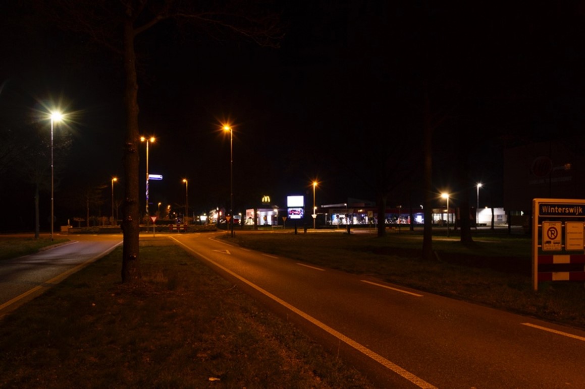 Straat met twee rijbanen, gescheiden door grasstrook. In het donker met straatlantaarns en in de verte andere verlichting en een rotonde. Zie bijschrift onder afbeelding